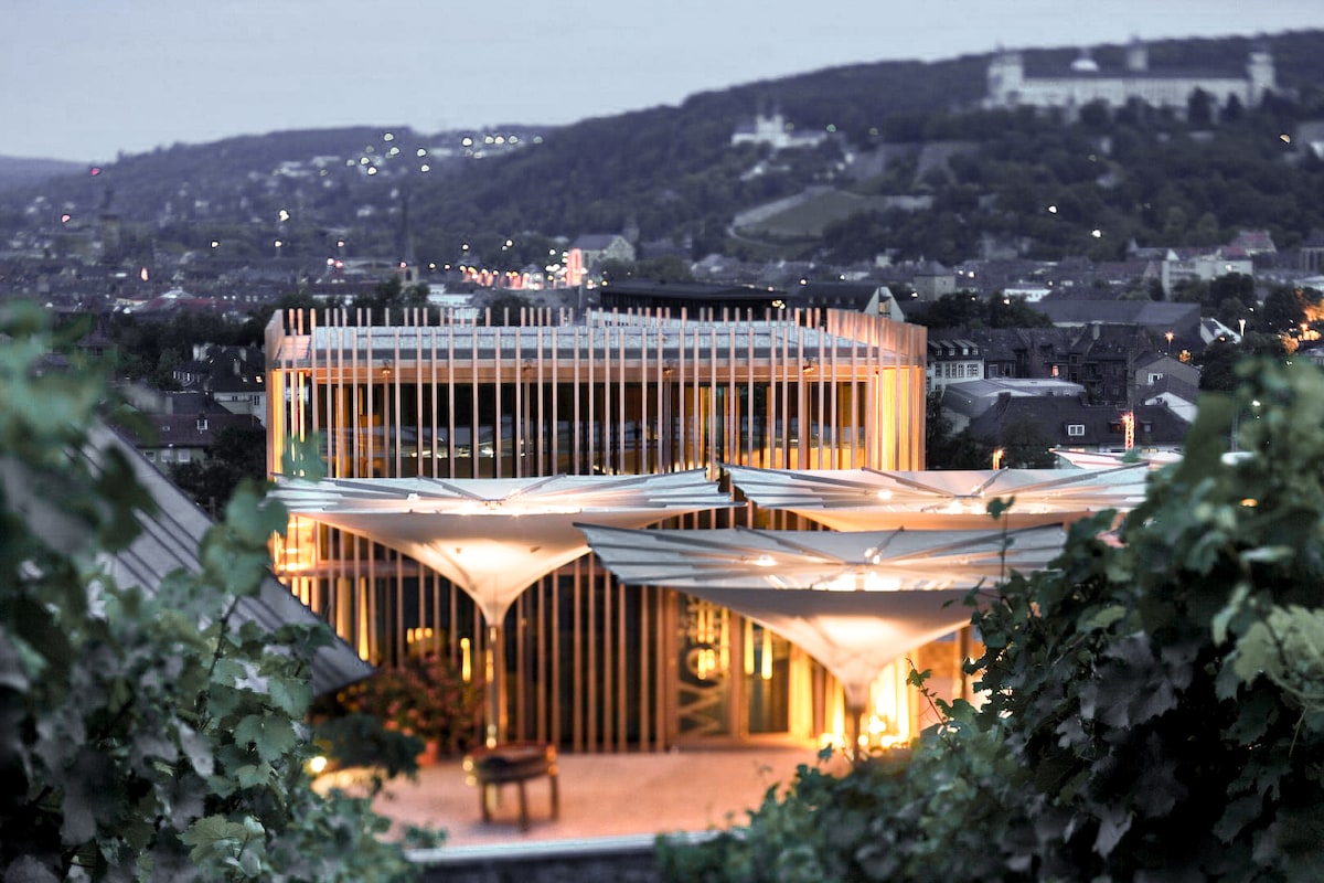 The exterior of the wine cellar features modern architectural elements, highlighted by illuminated wooden slats. Tensioned fabric canopies provide shade and aesthetic appeal. The surrounding vineyards enhance the serene setting, with the distant city skyline and hills softly glowing in the evening light.