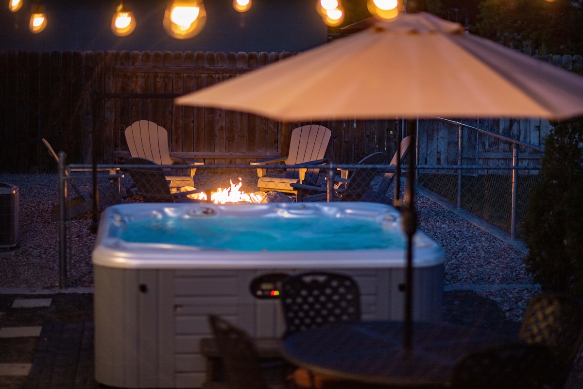 A hot tub occupies the foreground, with soft, ambient lighting from café-style lights above. Behind it, a fire pit with a flickering flame and seven Adirondack chairs creates an inviting outdoor space, surrounded by a fenced yard for privacy.