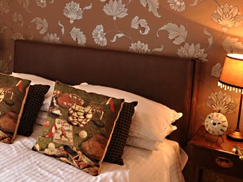 A cozy bedroom features a brown floral accent wall and a comfortable bed with plush pillows. A bedside table holds a classic lamp and a vintage clock, enhancing the room's inviting atmosphere. Warm lighting complements the decor, creating a tranquil space for relaxation.