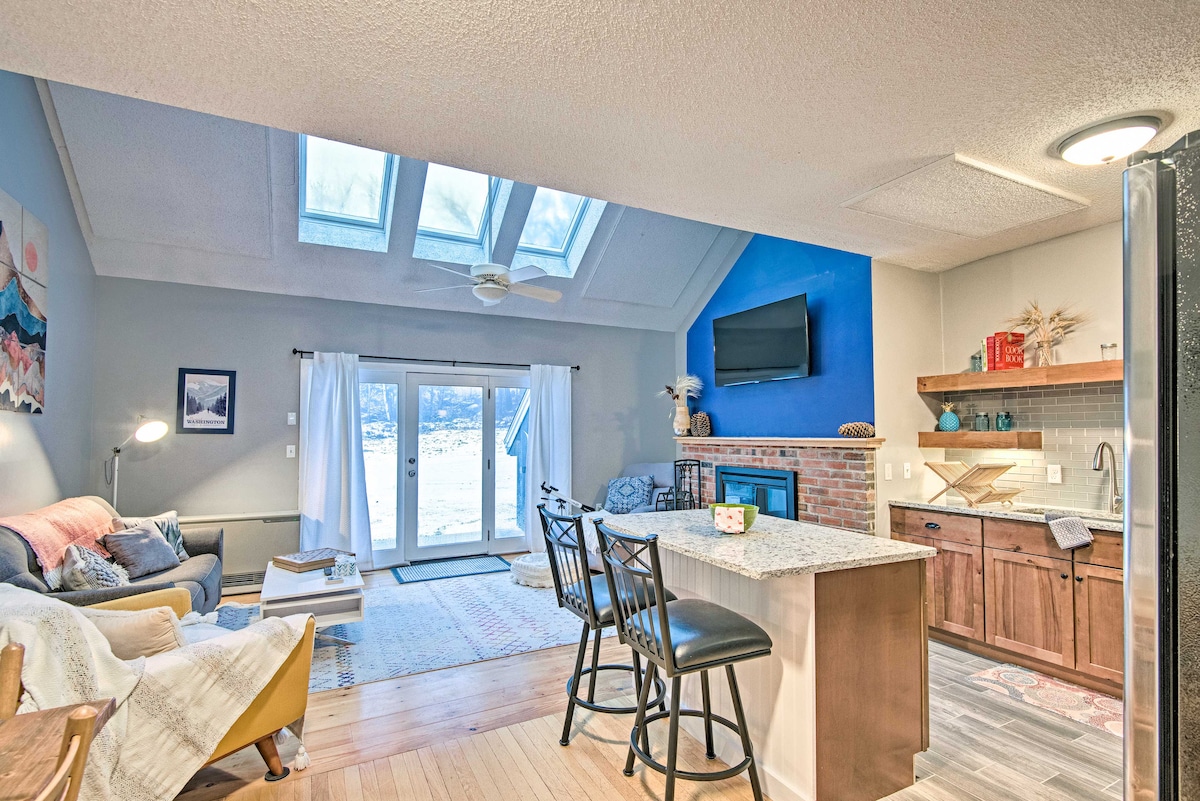 An open living area features large skylights allowing natural light to fill the space. A modern kitchen with a granite island and seating is visible alongside a comfortable sitting area, which includes a cozy sofa and soft furnishings. A brick fireplace and flat-screen TV add warmth and entertainment options.