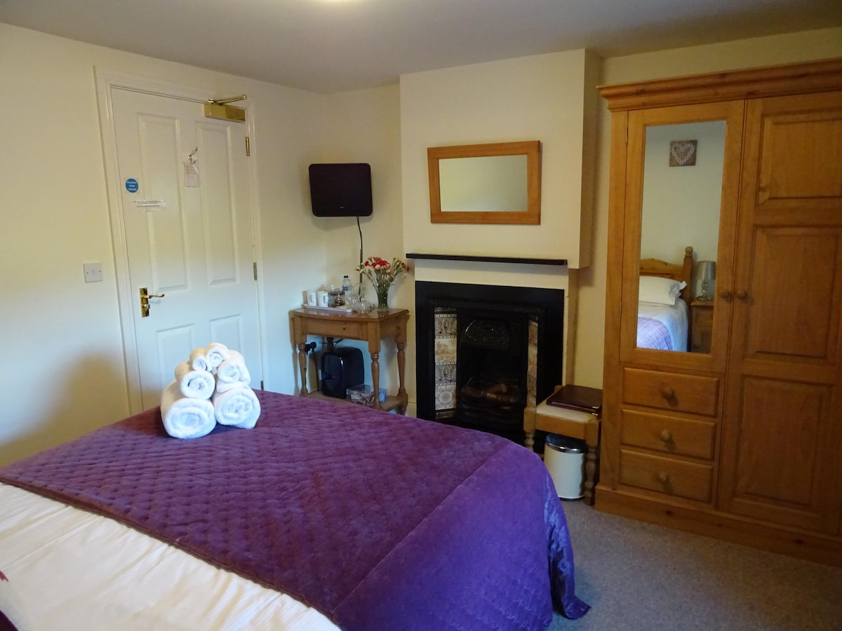 A cozy room features a kingsize bed covered with a deep purple quilt. A wooden wardrobe and mirror are present, along with a small TV mounted on the wall. White towels are neatly stacked on the bed, and a fireplace adds warmth to the atmosphere.