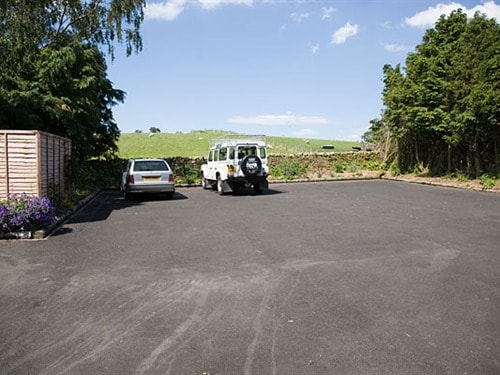 A spacious parking area is shown, featuring two parked vehicles. The surrounding landscape includes green hills, and a clear blue sky enhances the tranquil rural atmosphere. Decorative flowers can be seen alongside the parking space, contributing to the overall setting.