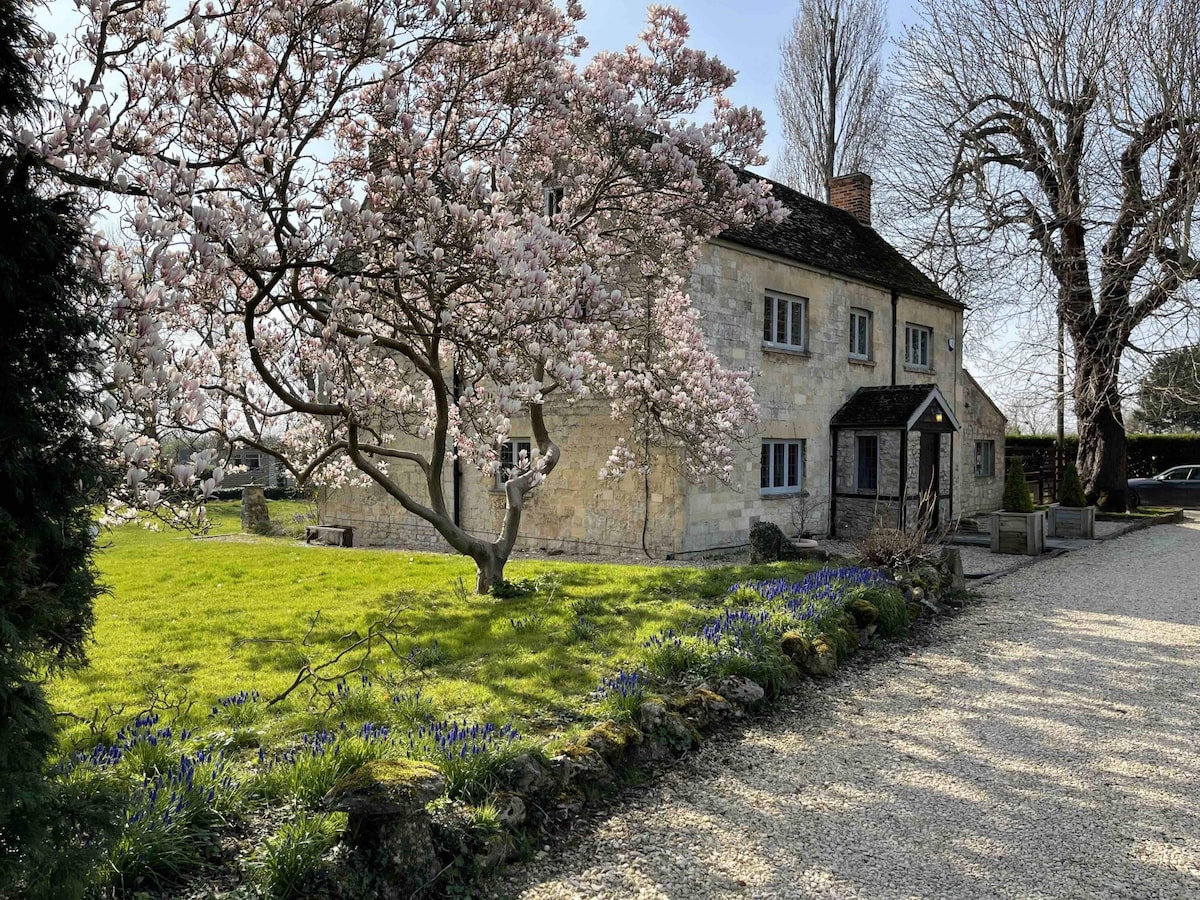 The exterior of the farmhouse is surrounded by lush greenery, highlighted by blooming magnolia trees and clusters of blue flowers. A gravel pathway leads to the private entrance, showcasing a blend of natural beauty and rustic architecture.