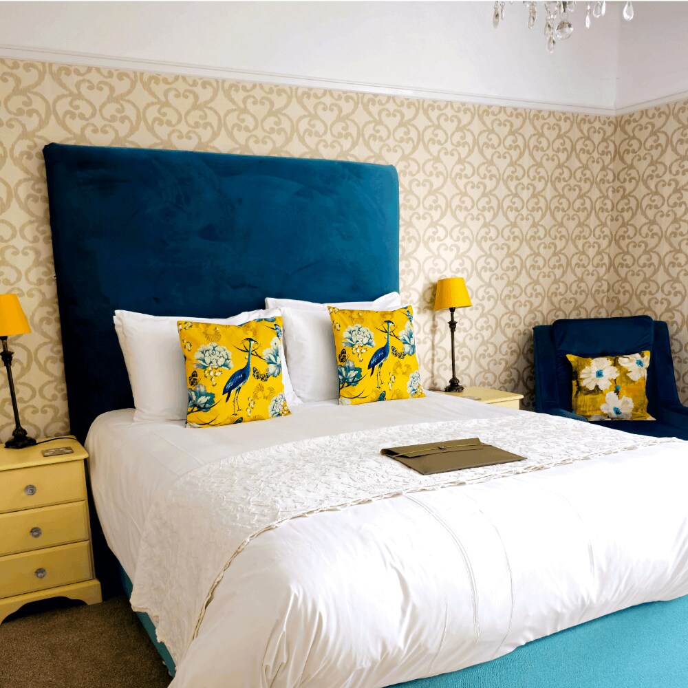 A bright bedroom features a spacious bed with a deep blue velvet headboard and patterned yellow accent pillows. Nightstands with lamps are positioned on either side, while a cozy chair sits in one corner, contributing to the room's inviting atmosphere.