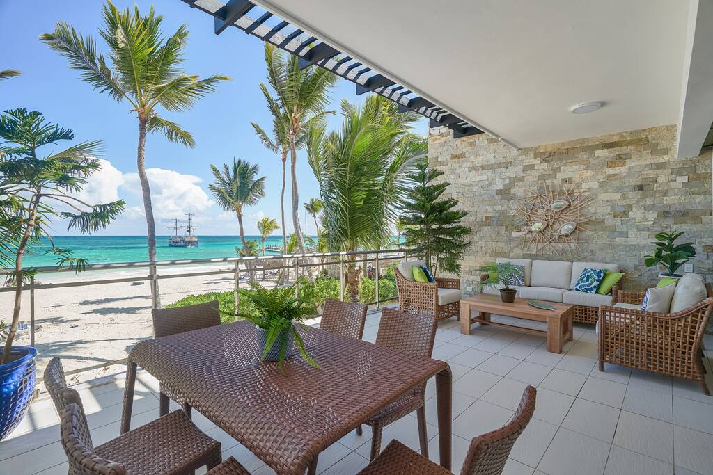 An outdoor balcony is depicted, featuring a dining table surrounded by six woven chairs. Coastal plants frame the space, and the view includes palm trees and the sandy beach beyond. Light-colored stone walls support the structure, with bright sunlight illuminating the comfortable seating area.