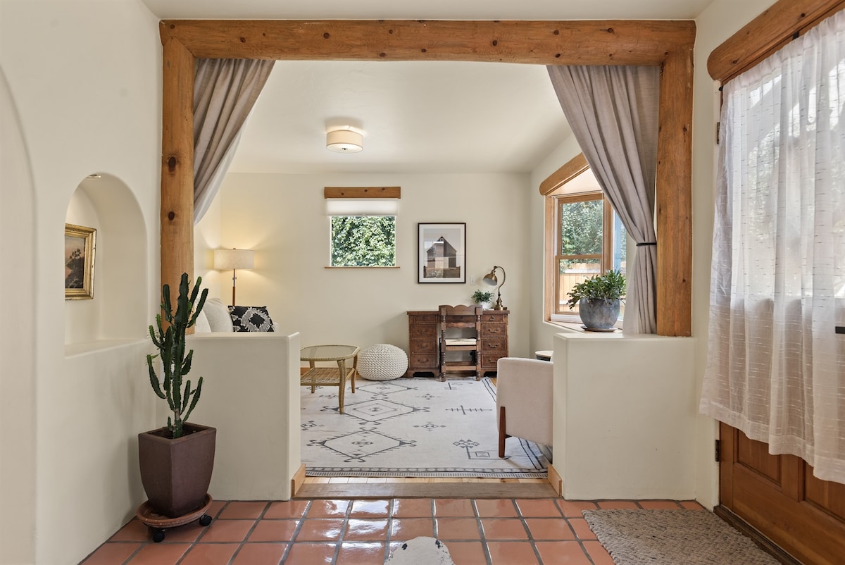 A welcoming sitting area features a light-filled ambiance with large windows. A cozy sofa and a wooden desk are positioned on a soft area rug, complemented by a potted cactus and a small white pouf. Soft curtains frame the view of the outdoors.