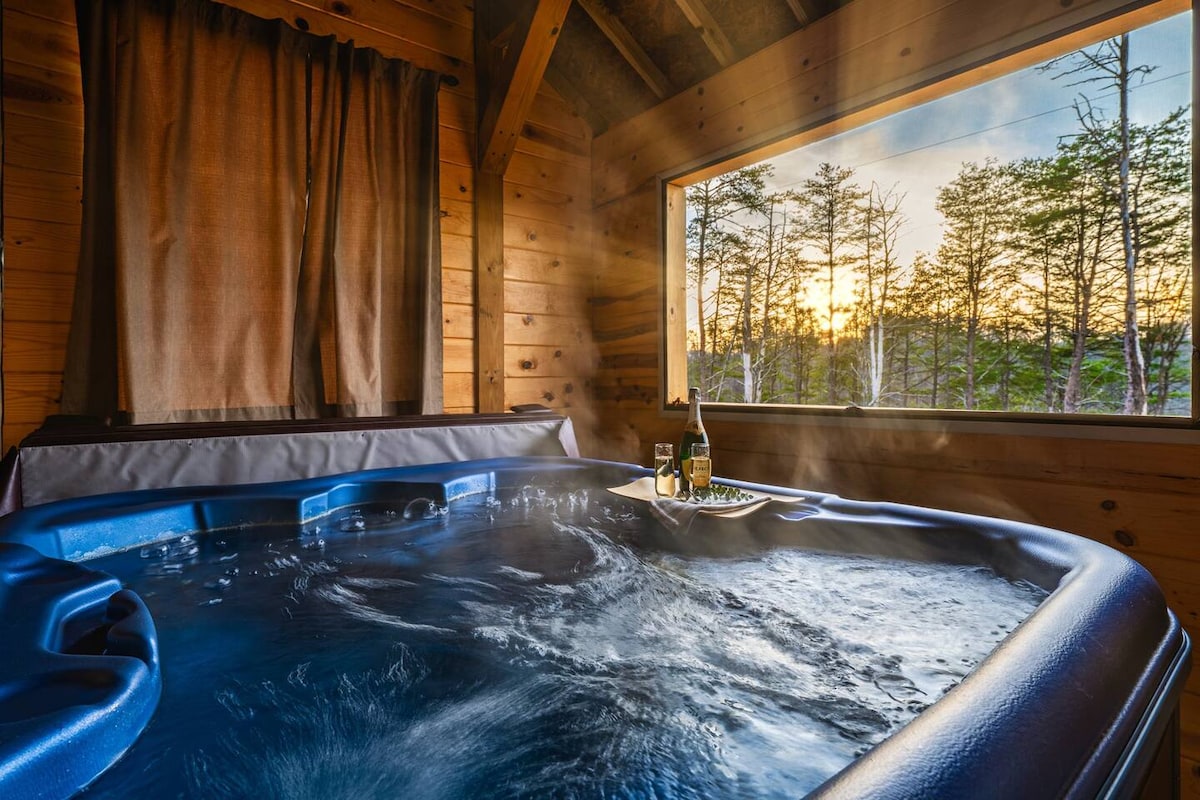 An indoor hot tub features swirling water, surrounded by wooden paneling. A tray with drinks rests beside the tub. Large windows allow natural light to filter in, revealing views of trees and a peaceful outdoor setting. Light fog enhances the tranquil atmosphere.