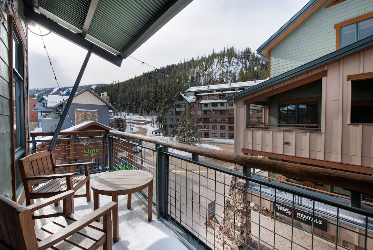 A balcony view features wooden chairs and a round table, overlooking a snowy landscape and nearby buildings. Pine trees border the scene, while distant mountains create a serene backdrop, providing a peaceful retreat from the bustle of the village.