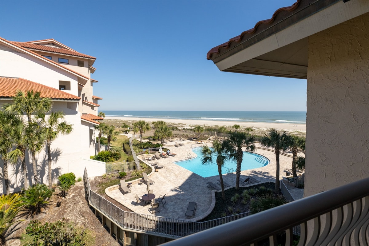 A view of the oceanfront pool area is visible from the spacious balcony. The pool is surrounded by lounge chairs and palm trees, with the beach and ocean in the background, under a clear blue sky.