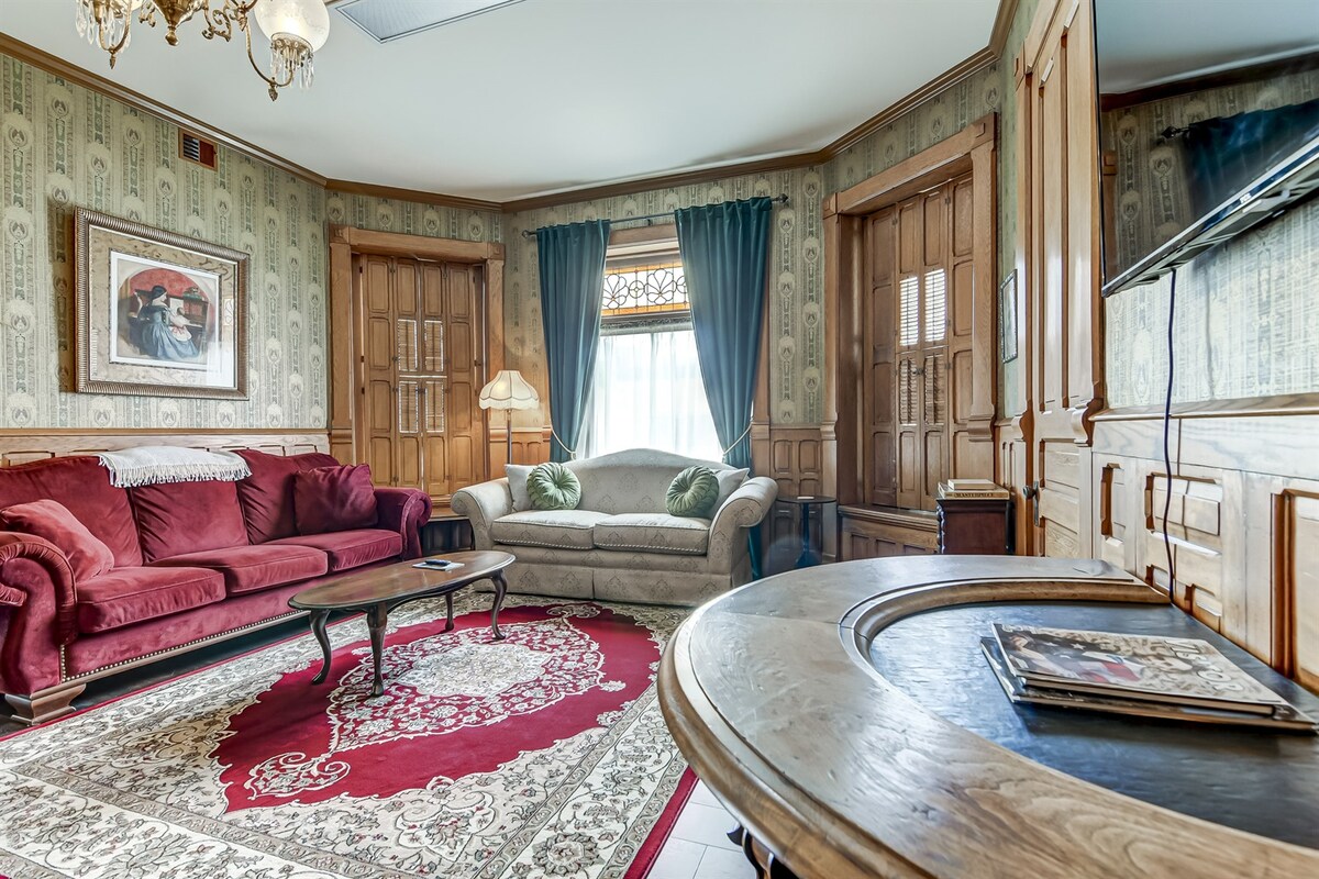A living room features a combination of red and beige furnishings, including a plush red sofa and a beige settee adorned with green cushions. Rich wood paneling frames the windows, which are draped in dark curtains. A traditional area rug anchors the space, with a circular table situated at the center.