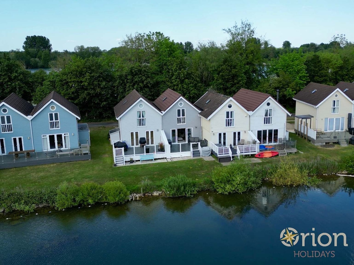 A row of New England style holiday cottages is positioned along the edge of a serene lake. Lush greenery frames the cottages, which feature large windows and decks overlooking the water. Each cottage exhibits a distinct color, adding charm to the lakeside setting.