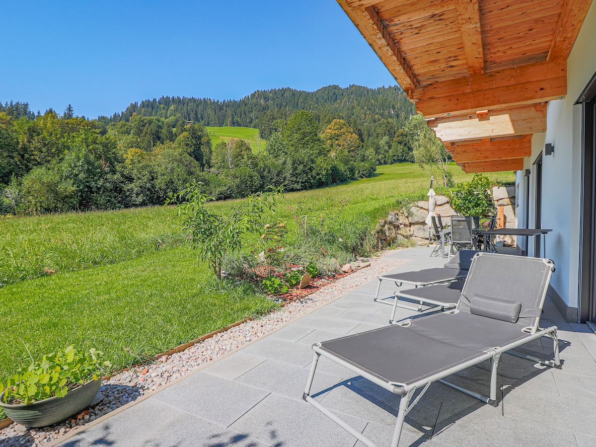 A spacious terrace is shown with two lounge chairs facing a lush green landscape. The view includes rolling hills and trees under a clear blue sky. Natural stone flooring leads to a well-maintained garden area with decorative plants.