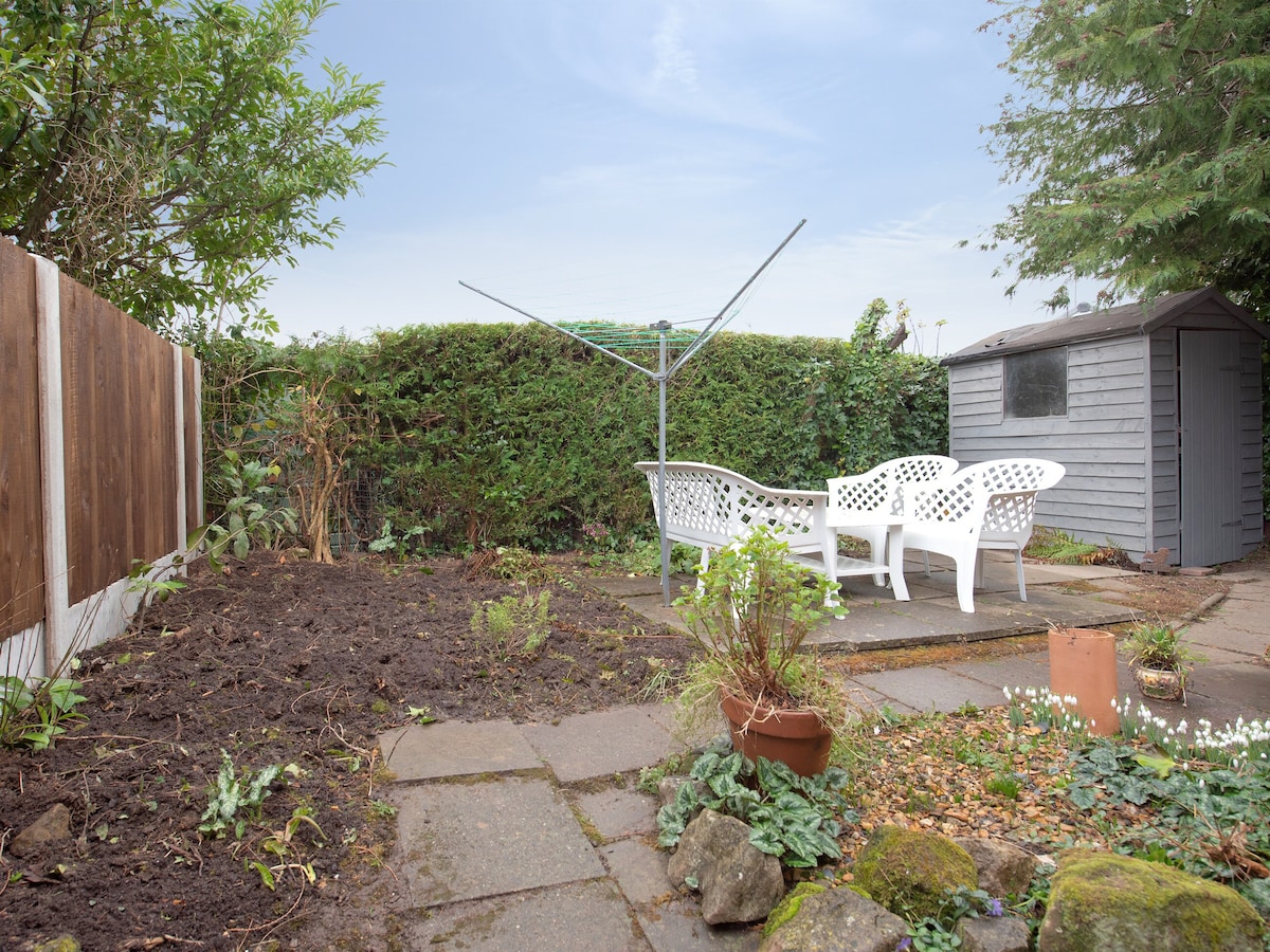 An enclosed back garden features a paved sitting area with white garden furniture, surrounded by greenery. A small shed is present in the background, while freshly turned soil and blooming plants are visible, creating a relaxing outdoor space.