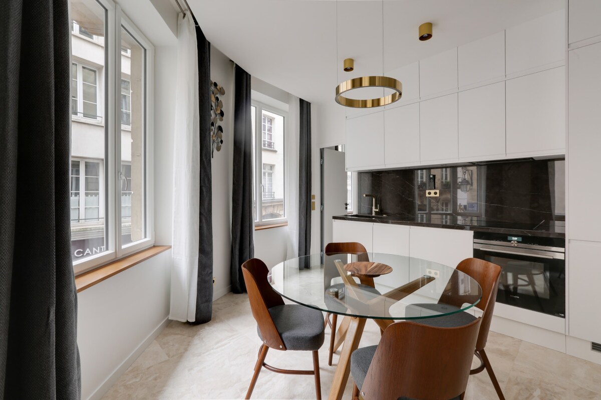 A modern dining area features a round glass table surrounded by wooden chairs. Large windows provide natural light, while neutral-colored curtains frame the view. The kitchen area is partially visible, showcasing sleek cabinetry and dark countertops.
