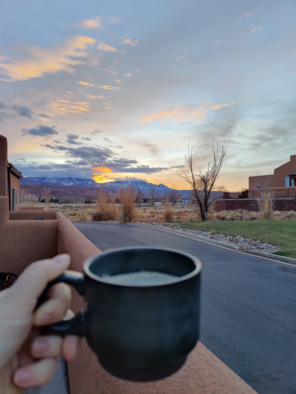 A hand holds a dark mug filled with a warm beverage, overlooking a quiet path that leads to a scenic view. The sky is painted with soft hues of orange and blue as the sun sets behind the mountains in the distance.