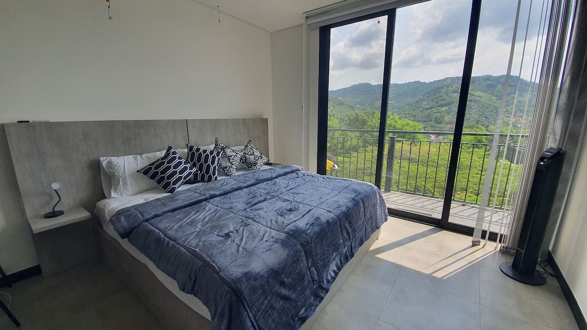 A spacious sleeping area is featured, with a large bed covered in a soft, textured blanket. Large glass doors open to a balcony, providing expansive views of green hills and natural surroundings. Natural light streams in, enhancing the serene environment.