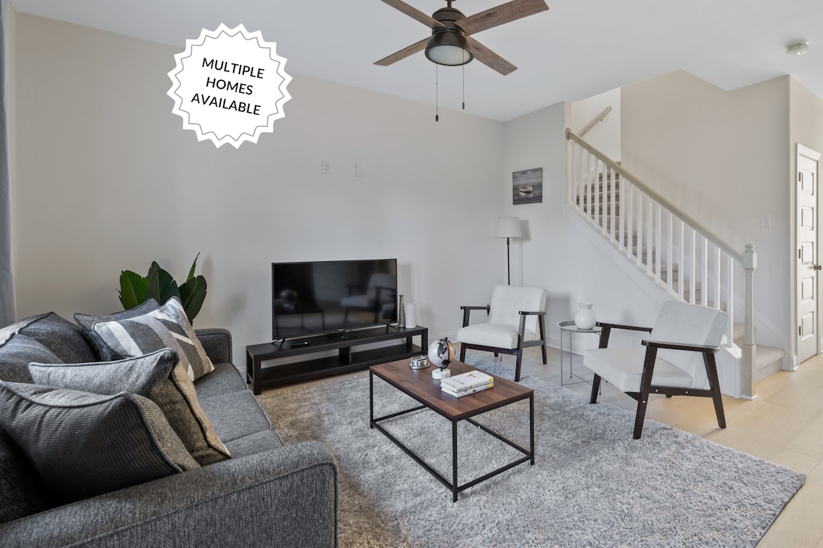 A modern living room is highlighted by a cozy grey sofa adorned with decorative pillows. Two armchairs complement a wooden coffee table at the center, while a 50” Smart TV is positioned against the wall. Natural light filters in, enhancing the inviting atmosphere.
