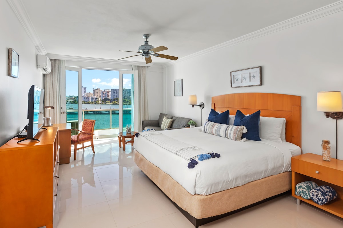A spacious studio features a king bed with decorative pillows, positioned across from a comfortable seating area. Large glass doors provide a view of the waterfront and city skyline, allowing natural light to fill the room. A flat-screen TV and wooden furnishings add to the cozy ambiance.