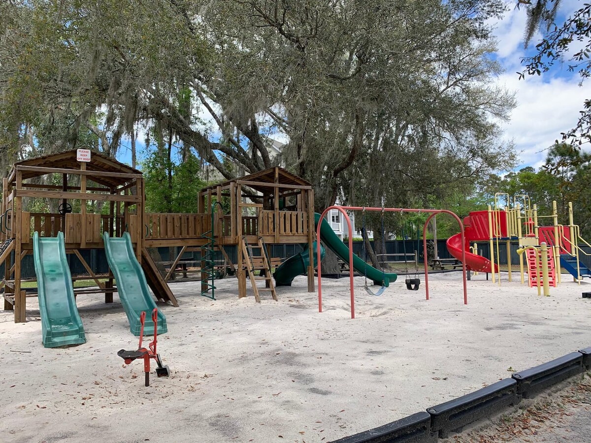 A playground area features a variety of play structures, including slides and climbing frames. Trees provide shade over the sandy ground, creating a safe and inviting space for children. Brightly colored equipment adds a cheerful touch to the outdoor setting.