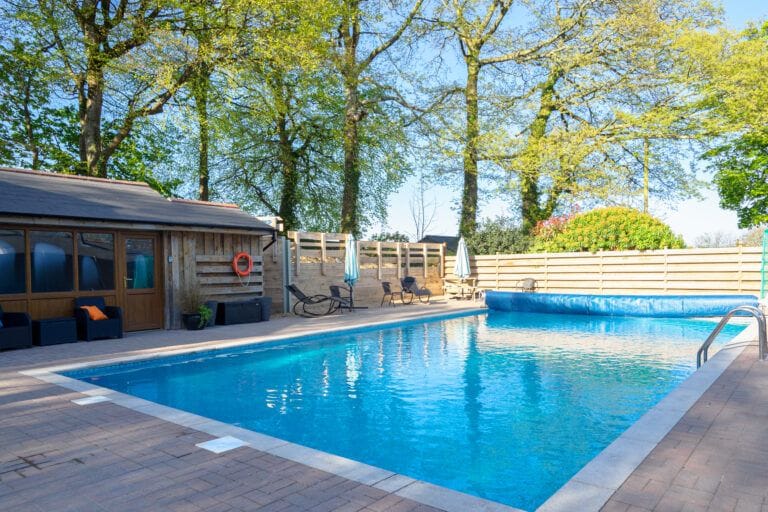 A large heated outdoor swimming pool is set within a landscaped area, surrounded by trees. Comfortable lounge chairs are positioned around the pool, and a wooden building with large windows is visible nearby. The water is clear and inviting, reflecting the blue sky above.