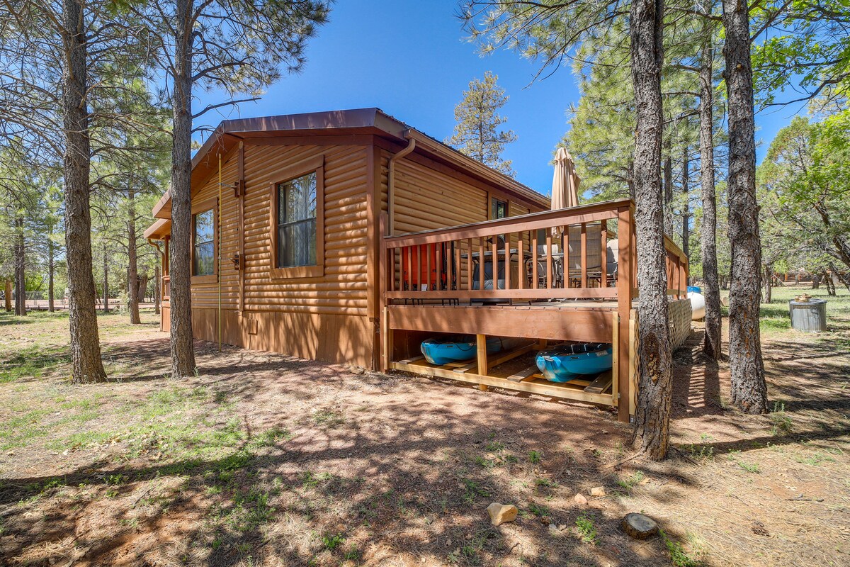 The cabin exterior features a wooden façade nestled among tall pine trees. A front deck with seating is visible, along with two kayaks stored below. Sunlight filters through the branches, enhancing the serene, natural setting.
