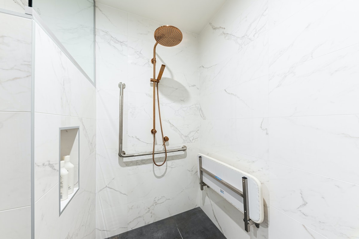 An accessible bathroom is shown, featuring a spacious walk-in shower with a rainfall showerhead mounted on a gold fixture. Grab bars are installed next to the shower for support, and a foldable grab seat is positioned against a white marble wall.