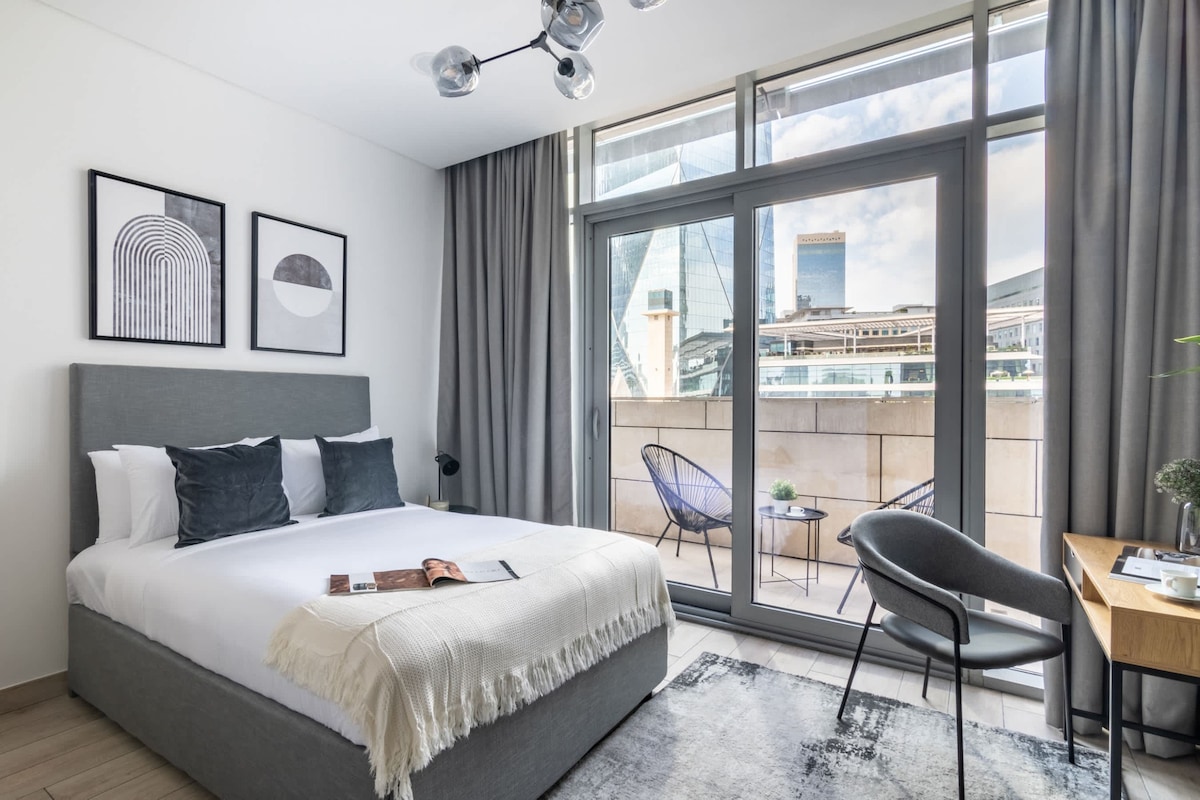 The studio features a spacious bed adorned with soft linens, complemented by modern art pieces on the walls. Natural light pours in through large glass doors that lead to a terrace. A functional workspace is provided, along with a cozy seating area for relaxation.