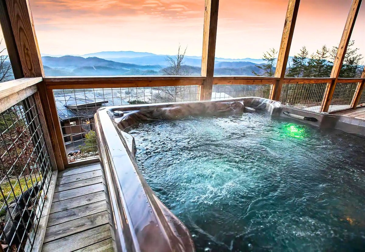 An outdoor hot tub sits on a wooden deck, surrounded by wooden railings. Scenic mountain views are visible in the background, with mist gently rising as the sun sets over the horizon, creating a serene atmosphere.