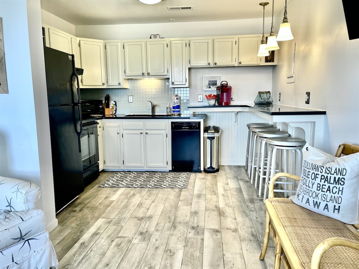 A well-equipped kitchen features white cabinetry and black appliances. Bar seating is provided at a counter with three metal stools. A patterned rug lies on the floor, and natural light fills the space from nearby windows, creating a bright and functional cooking area.