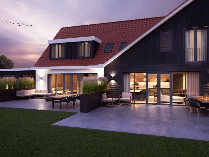A modern vacation home is showcased at dusk, featuring large glass doors that open onto a paved terrace. Comfortable seating is arranged outside, surrounded by green grass and planters. The sleek design includes a sloping roof, enhancing the spacious appearance of the property.