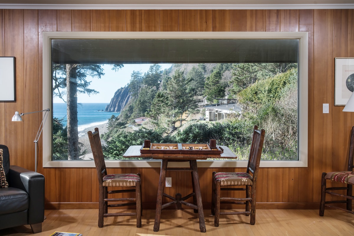 A large window offers stunning views of the ocean and surrounding greenery, creating a serene backdrop. A wooden table is paired with two matching chairs, inviting guests for a relaxing game or meal while enjoying the natural scenery.