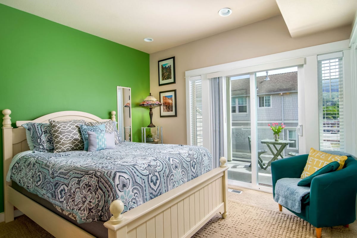A vibrant bedroom is highlighted by a green accent wall. A spacious bed is adorned with a patterned duvet, and an inviting chair is positioned beside a small table. Natural light floods the room through sliding glass doors, which lead to a private balcony with views.