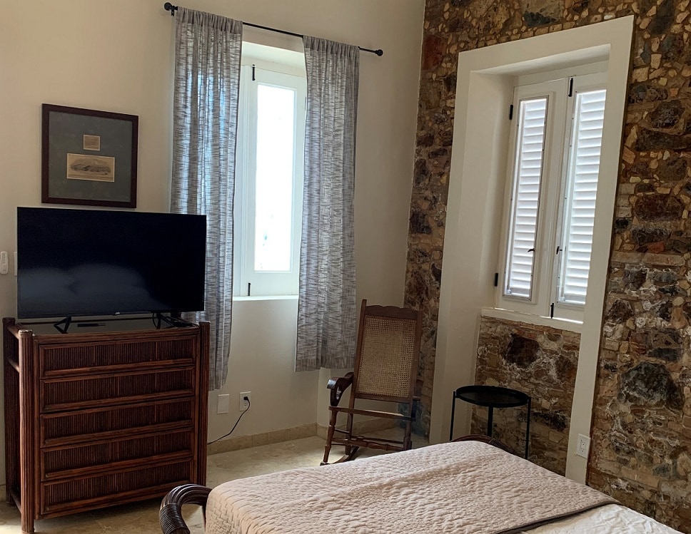 A guest room features rock-lined walls and large windows allowing natural light. A double-sized cradle bed is accompanied by a wooden dresser and a television. A rocking chair adds to the cozy atmosphere, while light curtains frame the windows.