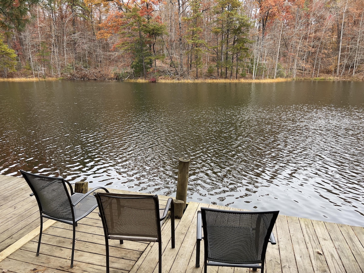 A wooden dock extends over the tranquil lake, featuring three black chairs positioned for seating. Surrounding trees display autumn foliage, reflecting in the calm water, enhancing the natural serenity of the lakeside setting.