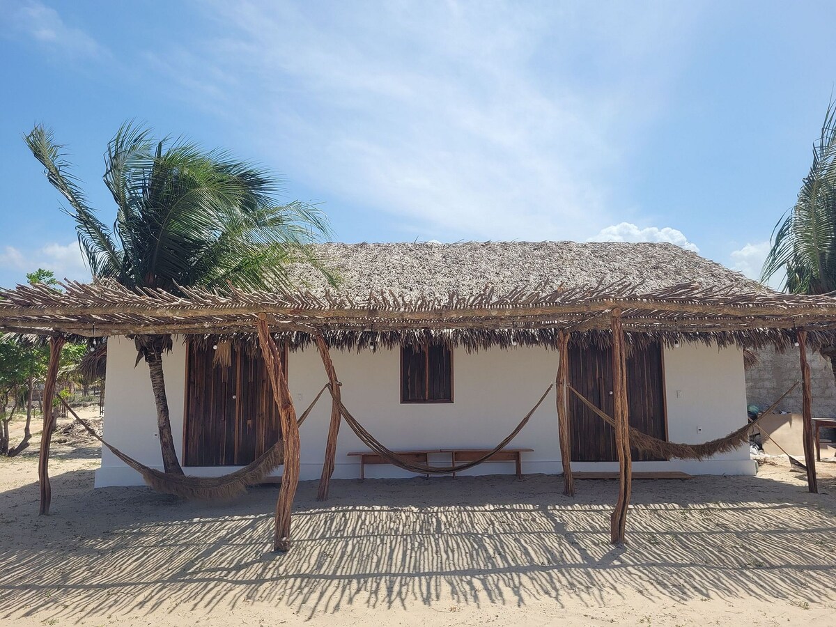 A simple structure featuring a thatched roof and wooden shutters is set against a clear blue sky. Two hammocks are suspended under a palapa, creating shaded lounging areas. The sandy ground is visible, providing a relaxed outdoor atmosphere.