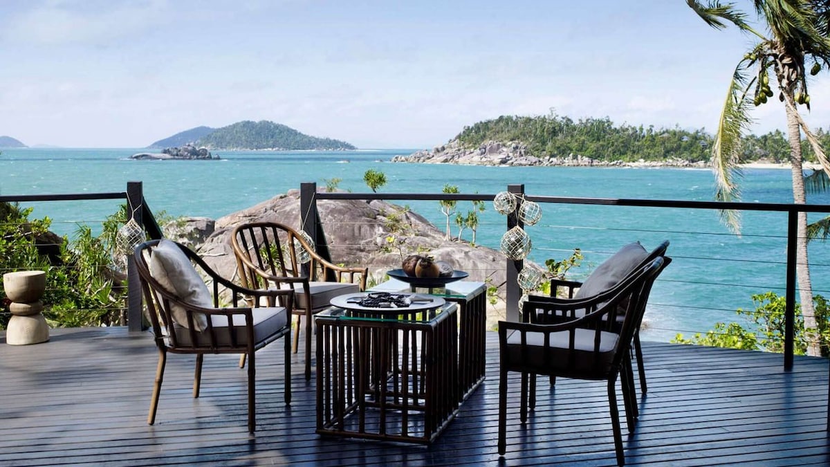 An outdoor seating area is situated on a wooden deck overlooking turquoise waters. Two armchairs and a round table are arranged for relaxation. Lush greenery and rocky island formations provide a stunning backdrop, enhancing the tranquil seaside ambiance.