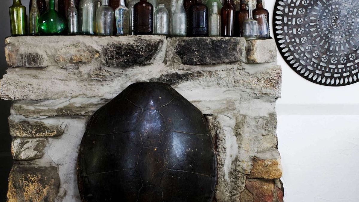 A stone fireplace features a unique black rounded design, resembling a turtle shell. Above, an arrangement of vintage glass bottles in various colors adds character, while a decorative circular wall piece in intricate pattern provides a striking contrast against the natural textures.