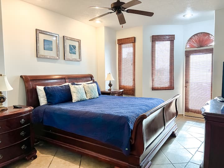 Примарна спална соба со брачен кревет, вентилатор на таванот, сопствена бања, ноќни шкафчиња и телевизор.