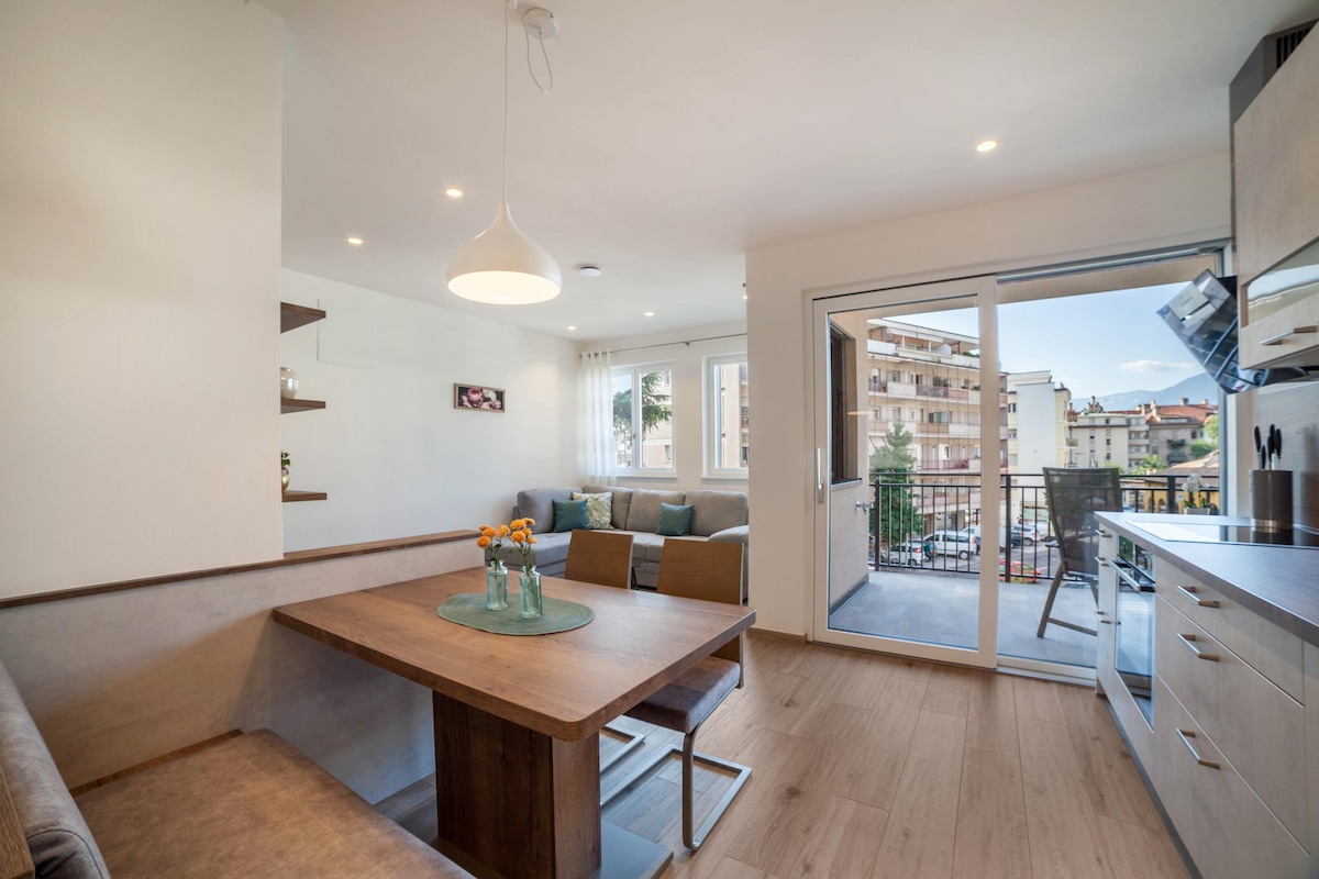 A modern living area features a dining table surrounded by chairs, with a comfortable sofa in the background. Large windows open to a balcony, allowing natural light to fill the space. Decorative shelves are mounted on the wall, adding a subtle touch of character.