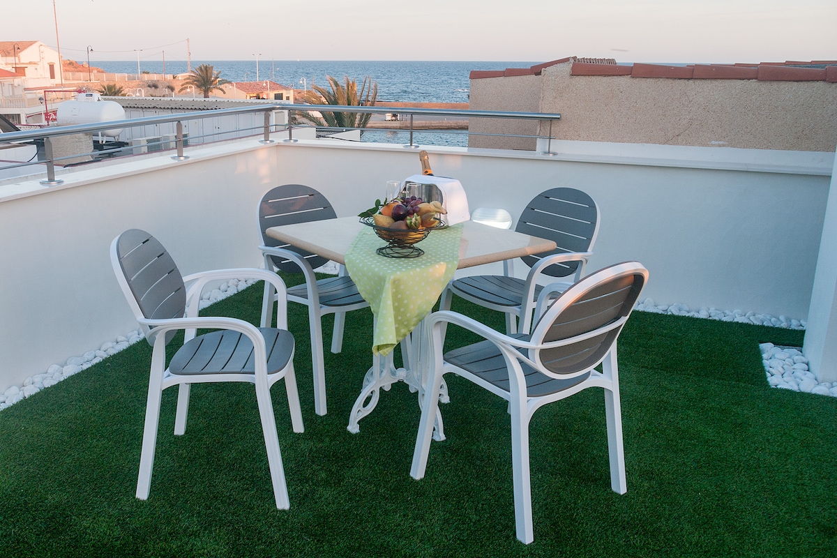 A balcony area features a table surrounded by four white chairs, set against a backdrop of the ocean visible in the distance. The table is adorned with a cloth and a basket of fruit, while artificial grass adds a green touch to the space.