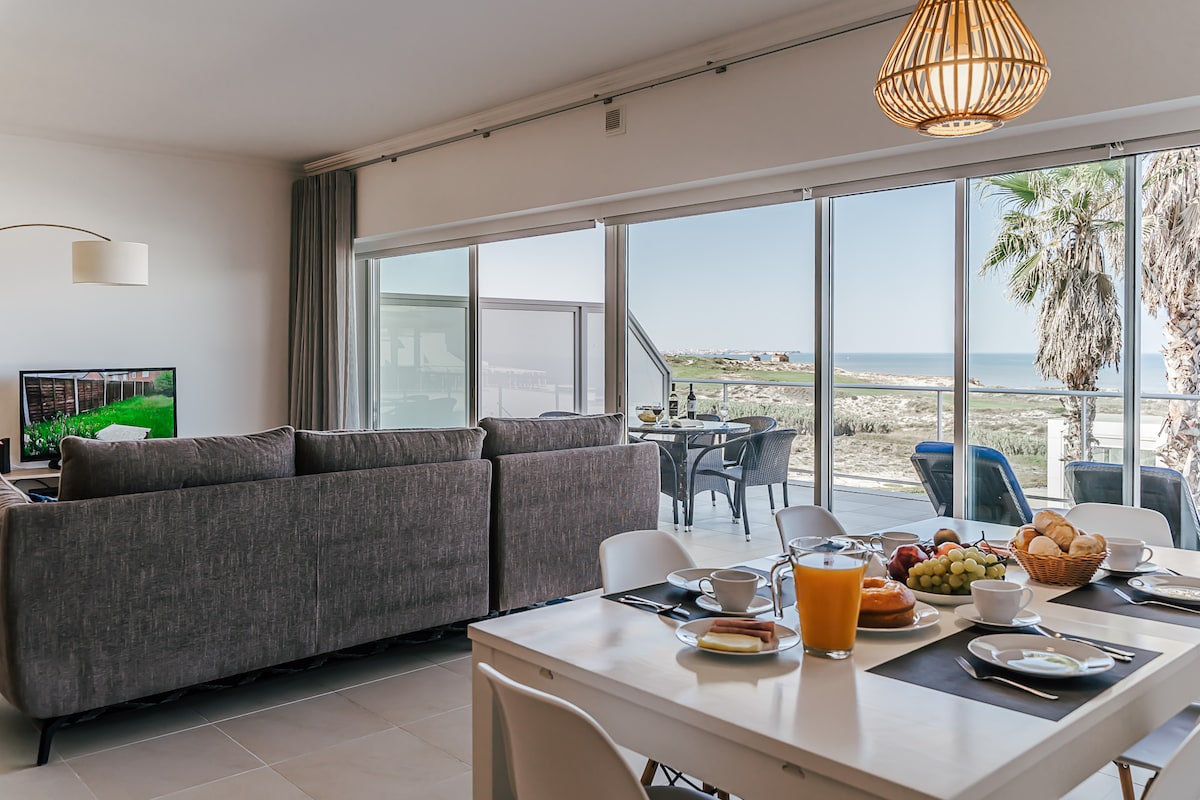 The dining area showcases a light wood table set with breakfast items, including fruit and pastries. A large sectional sofa faces a television, and expansive glass doors provide a view of the ocean and outdoor seating area. Natural light brightens the space.