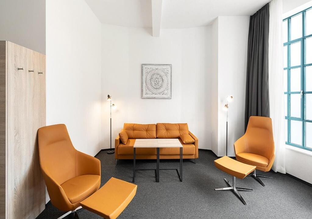 A modern seating area is displayed with a sleek orange sofa and two matching armchairs. A rectangular table is positioned in the center, complemented by stylish floor lamps. Large windows with dark curtains allow for natural light, enhancing the open and airy feel.