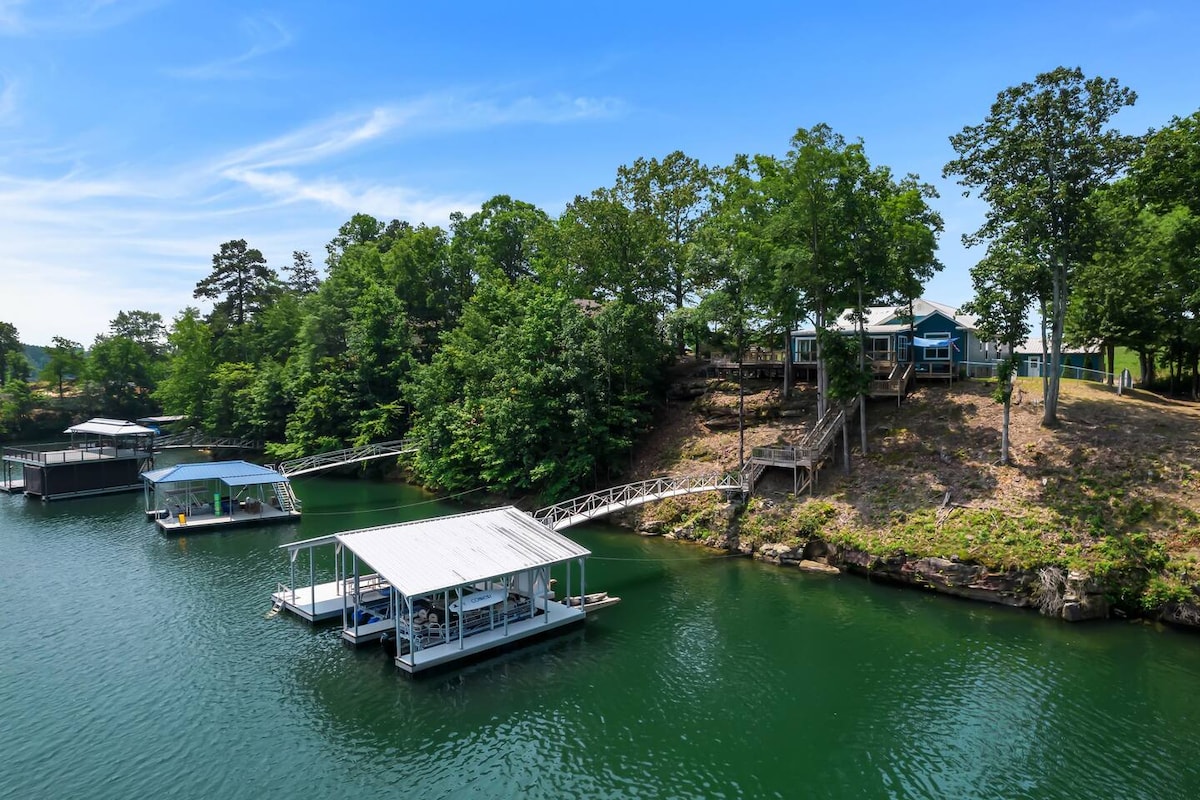 The serene lake setting features a calm, clear cove surrounded by lush greenery. Two boathouses with slips are positioned on the water's edge, alongside a wooden dock leading up to the property, which is nestled on a gently sloping bluff.