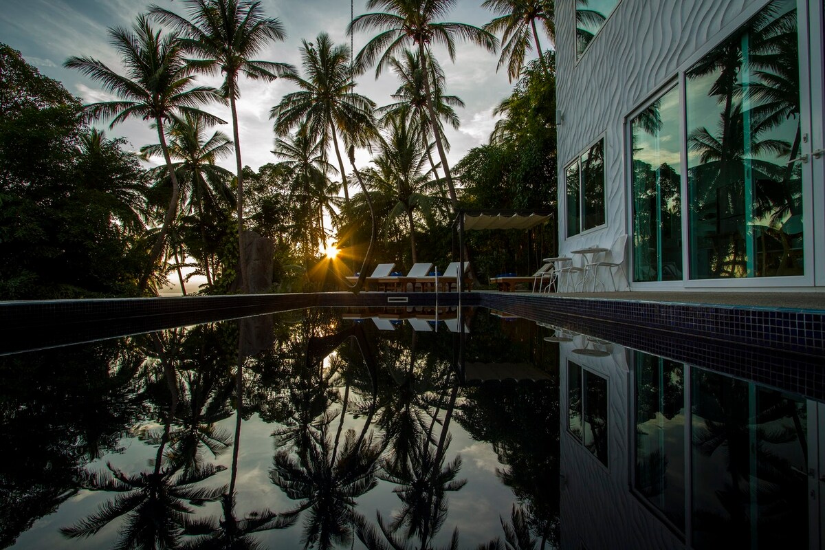 The tranquil private swimming pool reflects the surrounding palm trees and sunset, creating a serene atmosphere. Loungers are positioned nearby, with a shaded area visible in the background. Large windows of the villa provide a view of the lush greenery, enhancing the natural setting.