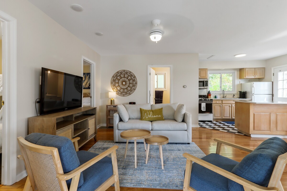 A welcoming living area is furnished with a light-colored sofa and two armchairs. A flat-screen television is mounted on the wall, while a round coffee table sits in front of the seating. Natural light enters through nearby windows, highlighting the warm wood flooring.
