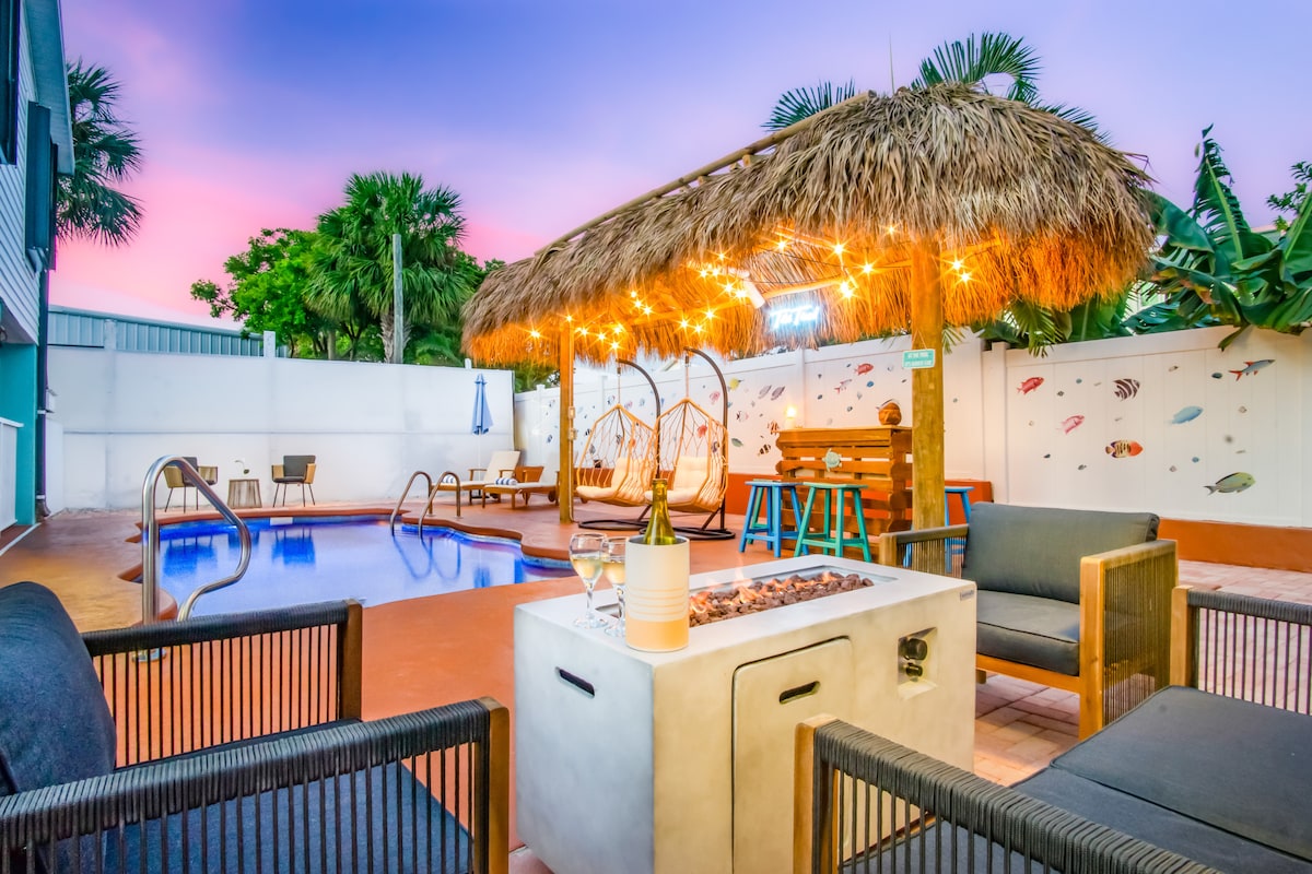 A tropical backyard features a private saltwater pool surrounded by seating areas. A thatched-roof Tiki Hut is adorned with string lights. A gas fire pit sits centrally, while walls are embellished with colorful fish murals. Lush greenery completes this inviting outdoor space.