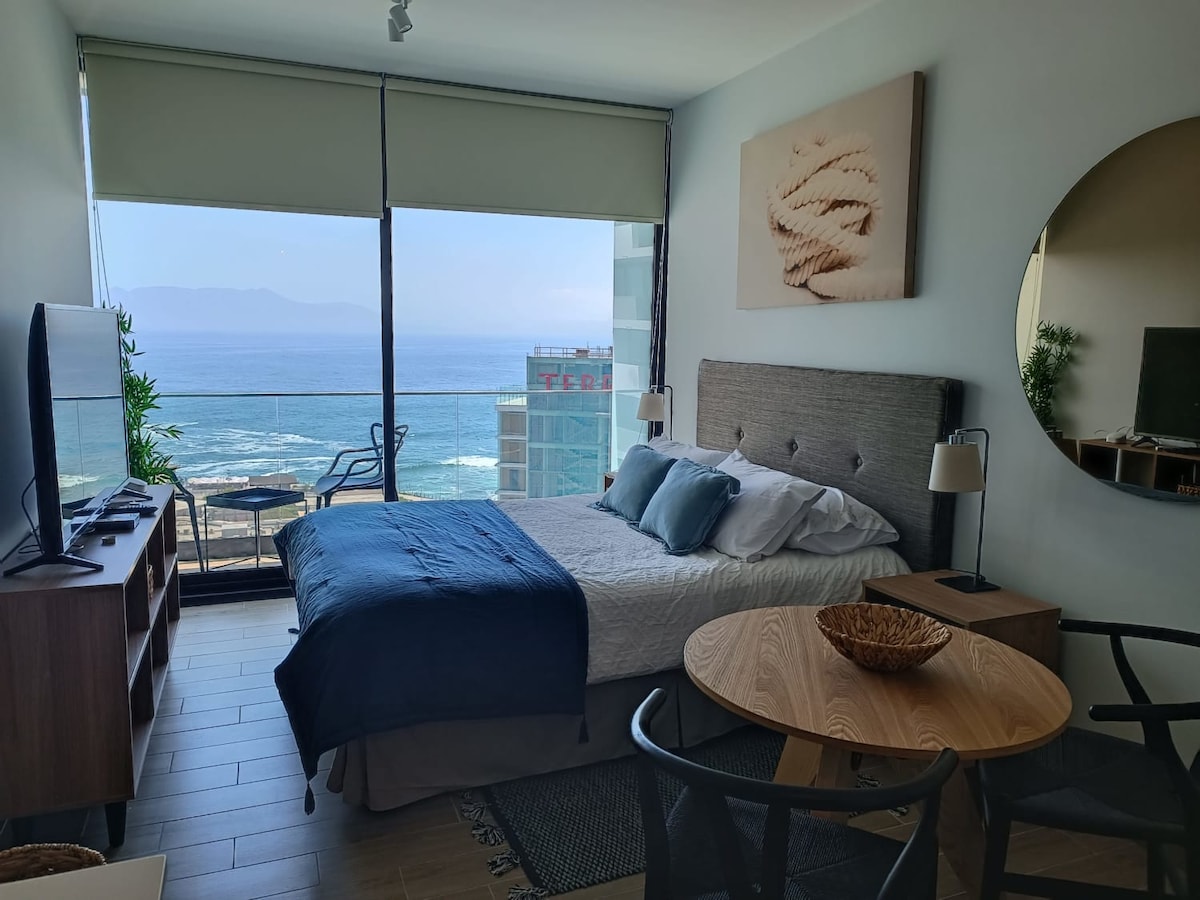 A bright and airy studio features a spacious bed adorned with soft linens, positioned next to large windows offering panoramic views of the ocean and city. A comfortable seating area is visible, accompanied by a round dining table, providing a cozy space to relax or enjoy meals.