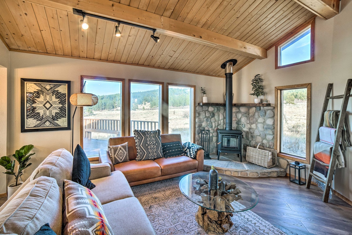A spacious living area features large windows that provide views of the outdoor landscape. A wood-burning furnace is positioned beside a stone fireplace. A comfortable sectional sofa and a round glass coffee table contribute to the inviting atmosphere, enhanced by warm wood accents on the ceiling.