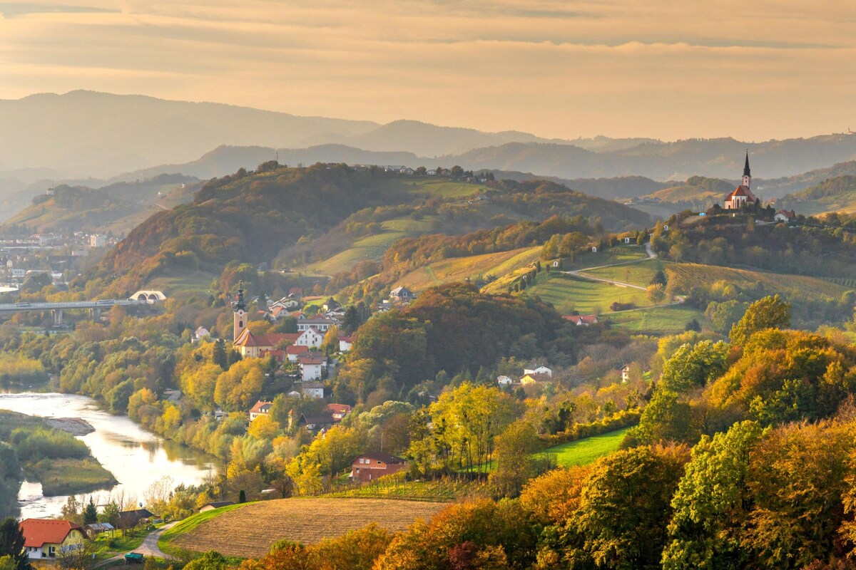 A panoramic view of rolling hills and vineyards extends into the distance, highlighting a small village nestled among greenery. A river meanders through the landscape, with soft sunlight casting a warm glow over the scene. The silhouette of a church steeple stands out against the backdrop of hills.