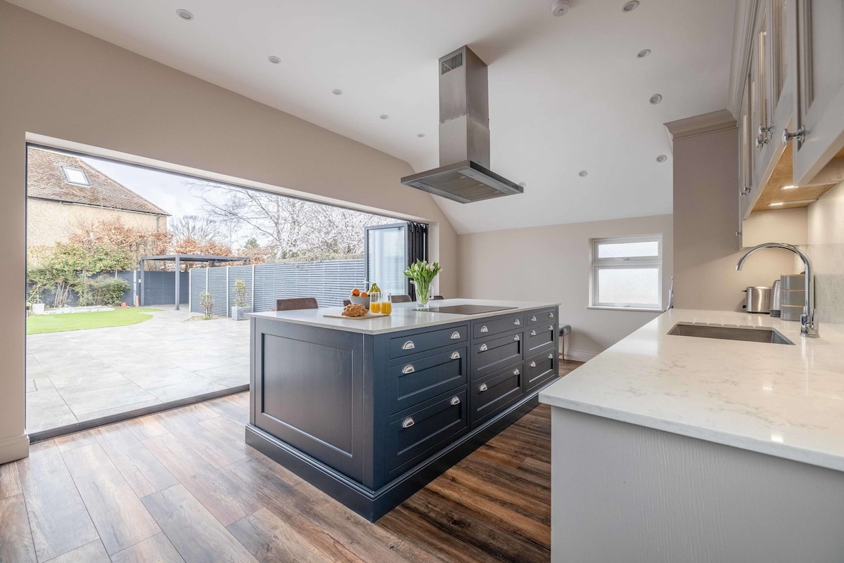 A spacious kitchen features a large central island with a dark blue finish, complemented by modern cabinetry and countertops. Bi-fold doors open to the private garden, allowing natural light to fill the room. Subtle decor elements add a contemporary touch.